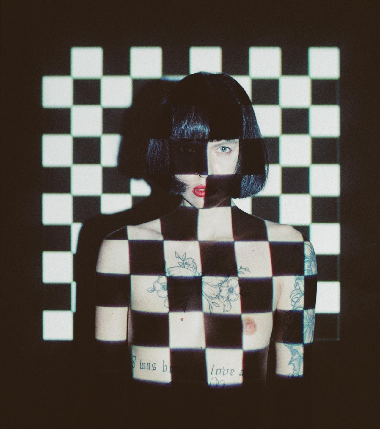 A woman with short dark hair and red lipstick is covered in a black and white checkerboard pattern projected onto her body and the wall behind her.