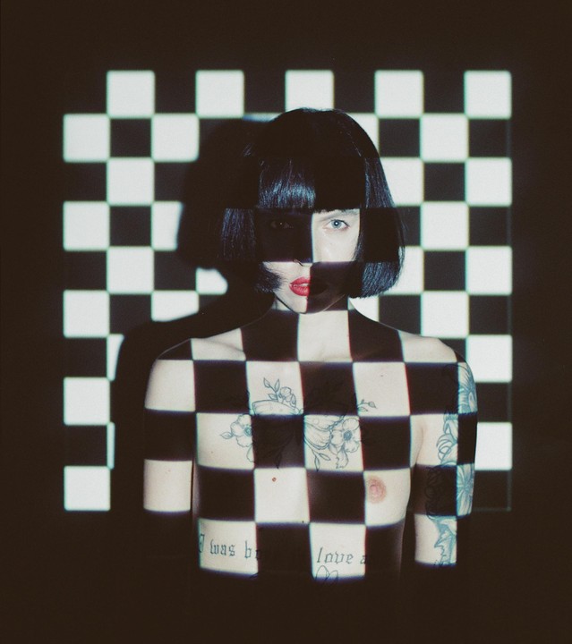 A woman with short dark hair and red lipstick is covered in a black and white checkerboard pattern projected onto her body and the wall behind her.