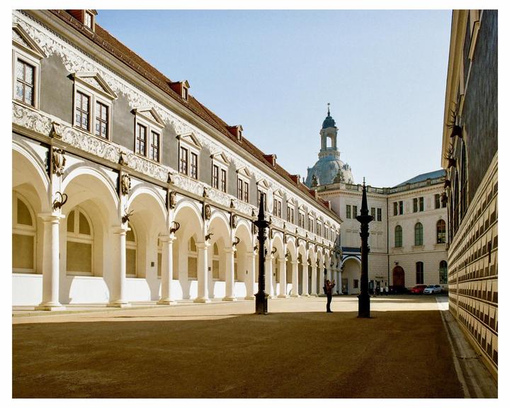 A person stands in a sunlit courtyard surrounded by ornate buildings with arched walkways and a distant dome.
