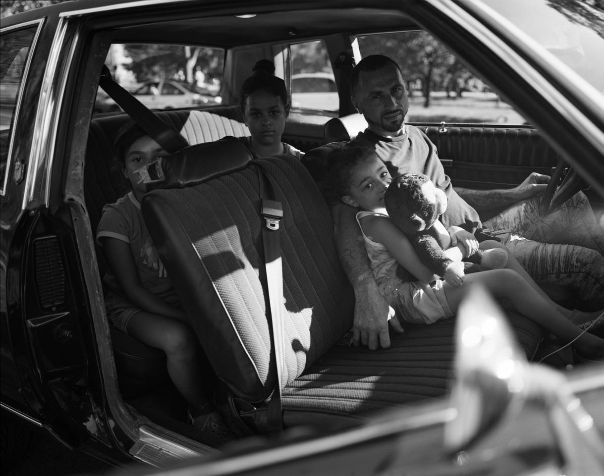 A man sits in the driver's seat of a car with three children, one of whom holds a stuffed animal.