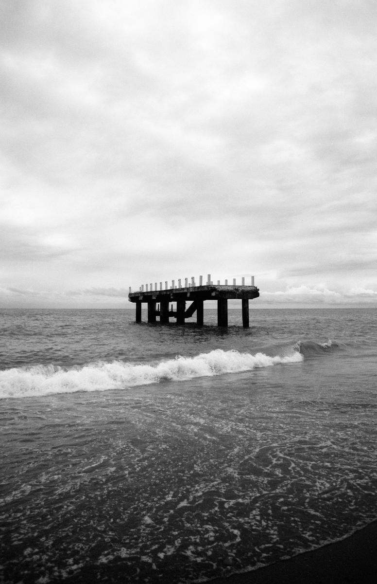 A concrete pier stands in the ocean under a cloudy sky, with a wave breaking in the foreground.
