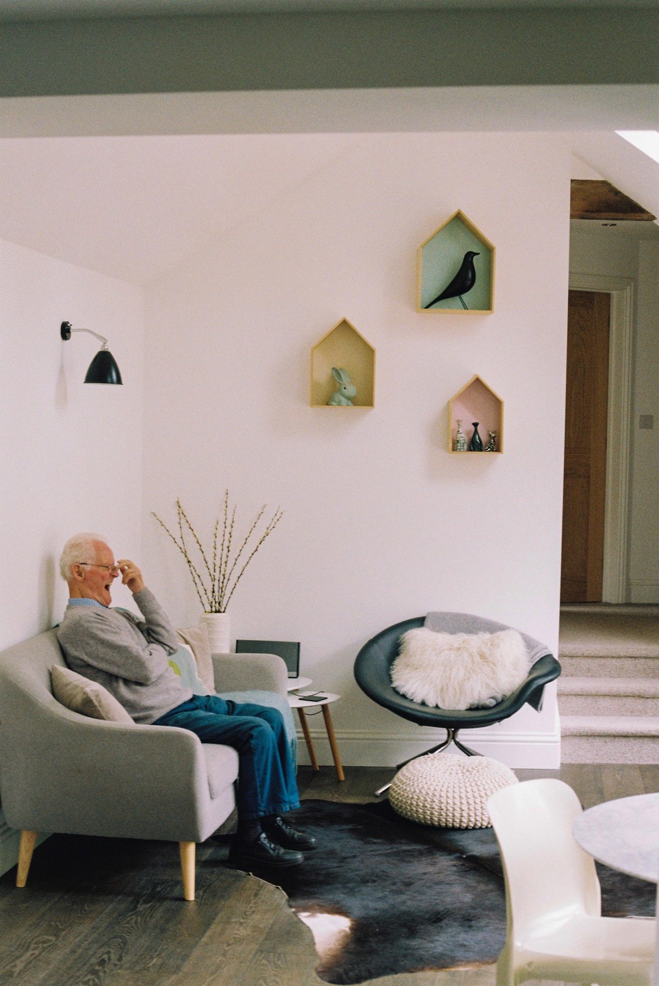 An older man sits on a sofa in a living room, laughing with his hand to his face.