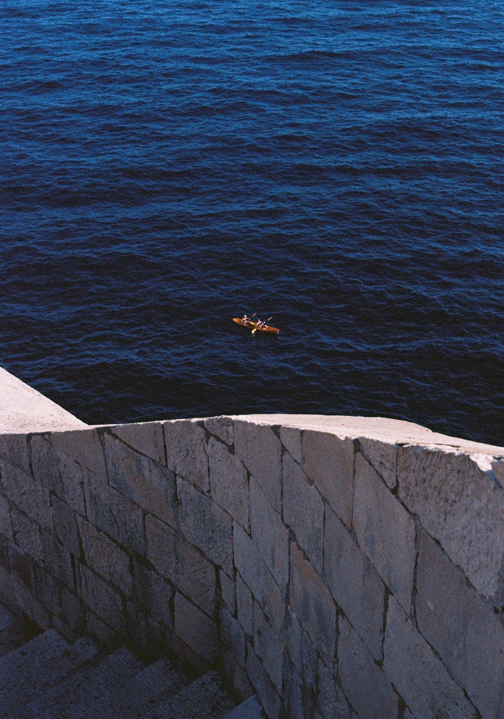 Two people in a kayak paddle on dark blue water next to a stone wall and stairs.