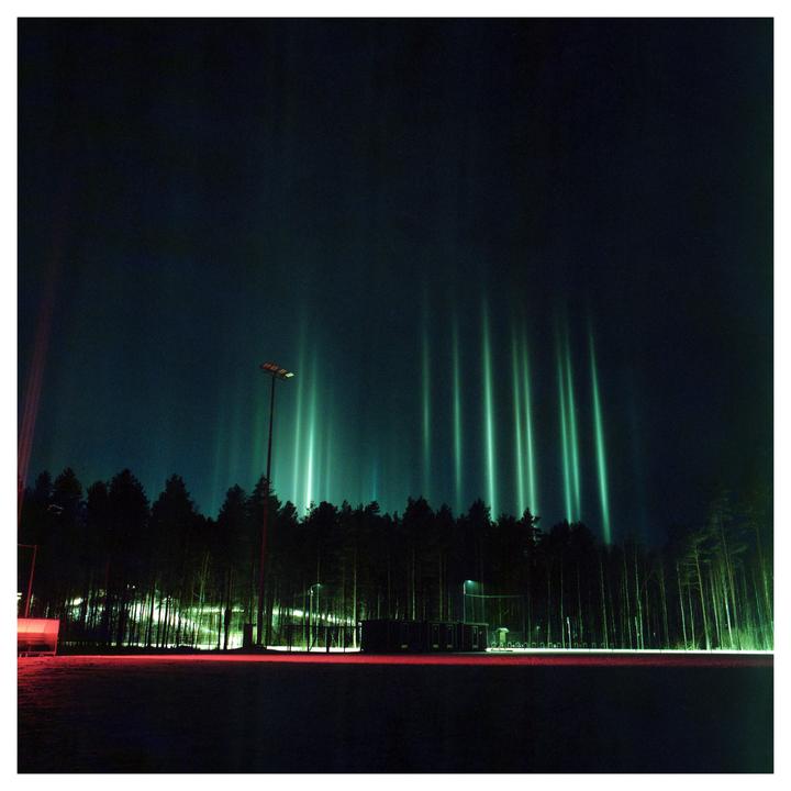 Green light pillars rise above a dark forest at night, with a red light streak in the foreground.