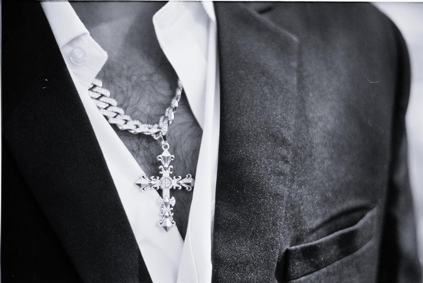 A man wears a cross pendant on a chain over a white shirt and under a dark suit jacket.