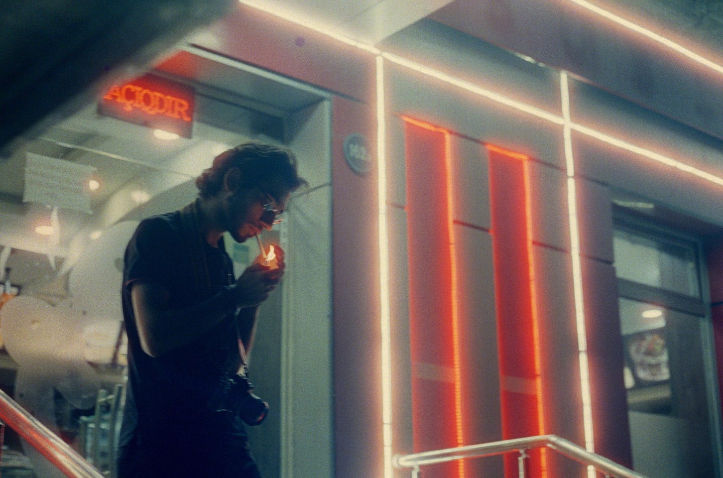 A man with a camera around his neck lights a cigarette at night in front of a building with neon lights.