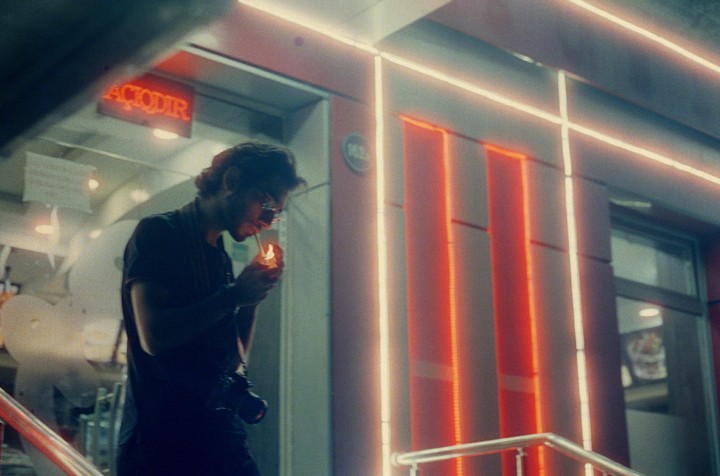 A man with a camera around his neck lights a cigarette at night in front of a building with neon lights.