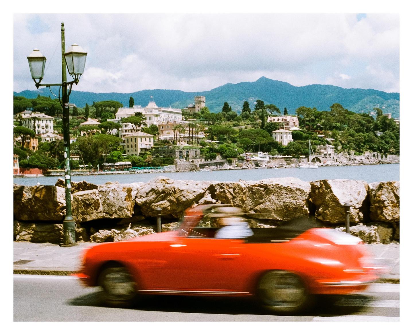 A red convertible car drives past a stone wall with a town and mountains in the background.