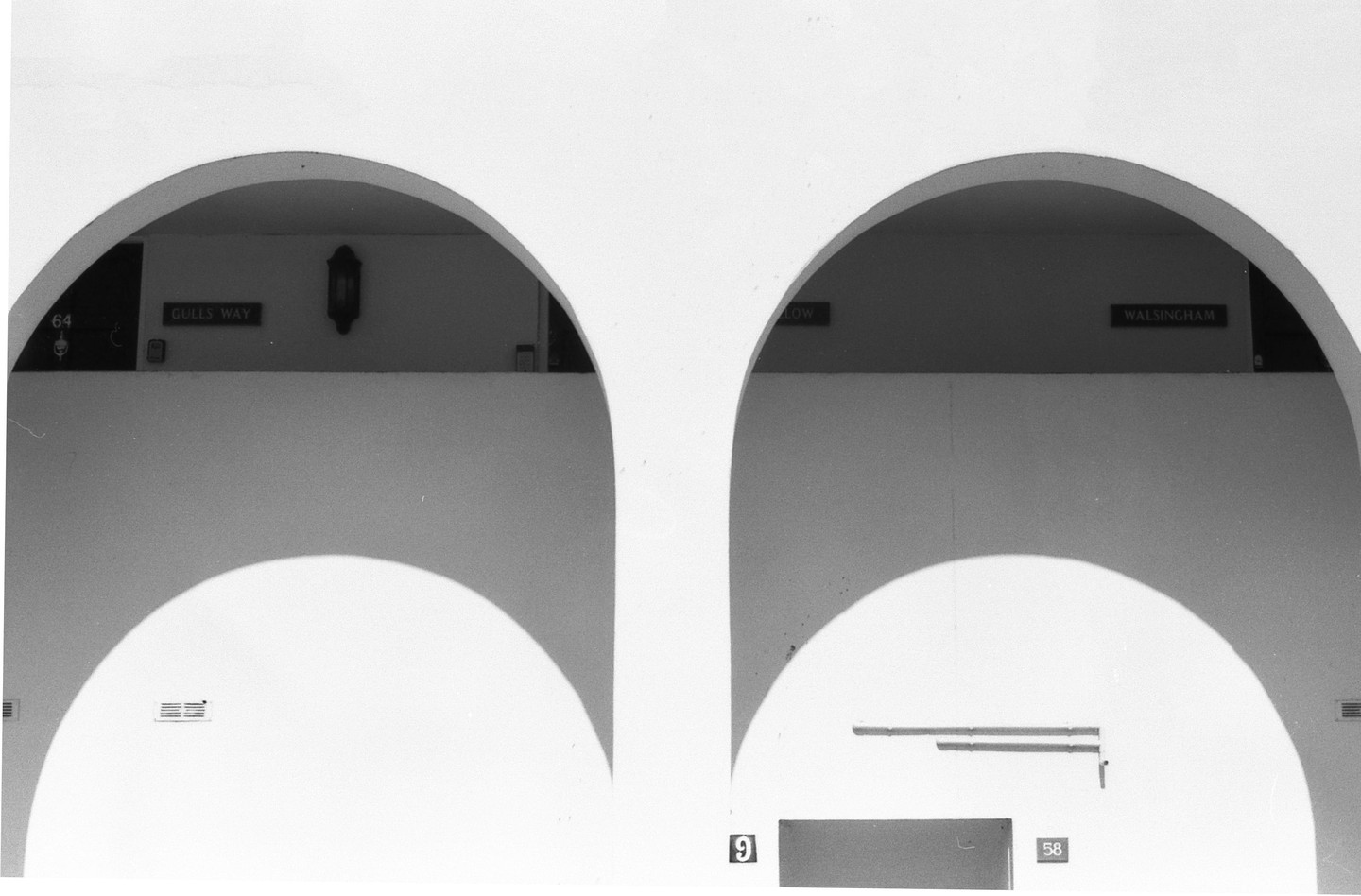 Two white arches cast shadows on a wall with signs for "GULLS WAY" and "WALSINGHAM" visible through them.
