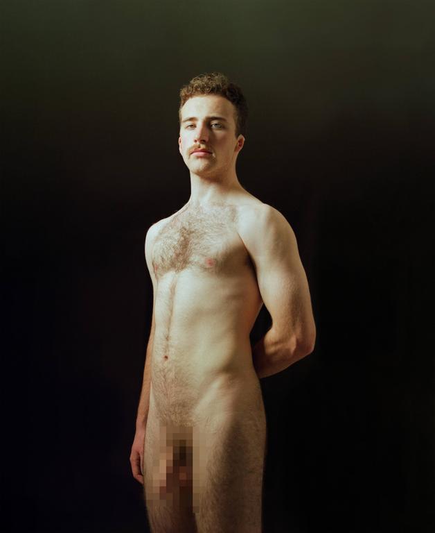 A man with a mustache stands nude against a dark background, looking directly at the viewer.