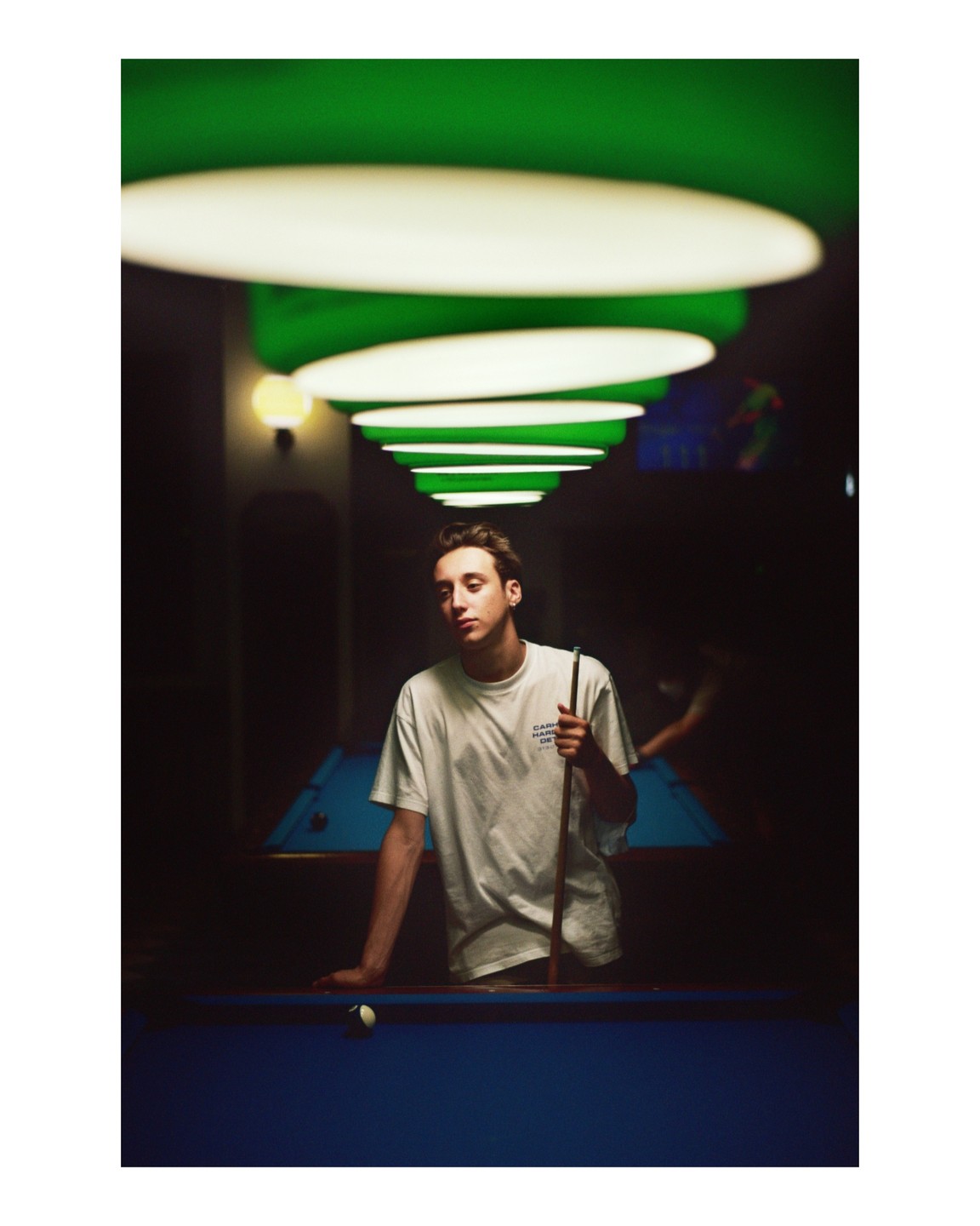 A young man stands next to a pool table, holding a cue stick, under a row of green and white lights.