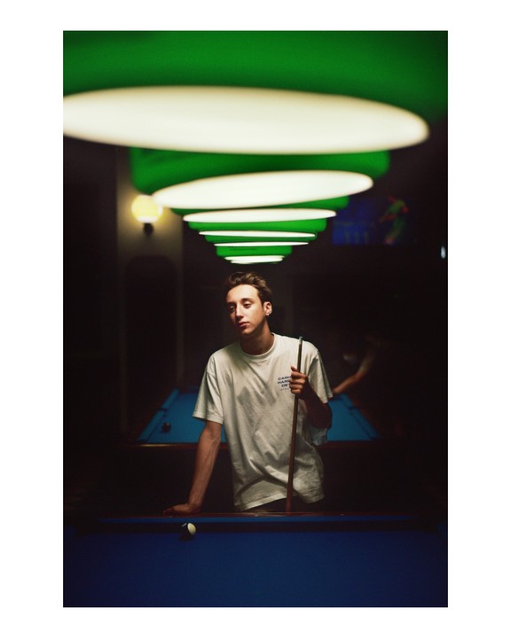 A young man stands next to a pool table, holding a cue stick, under a row of green and white lights.
