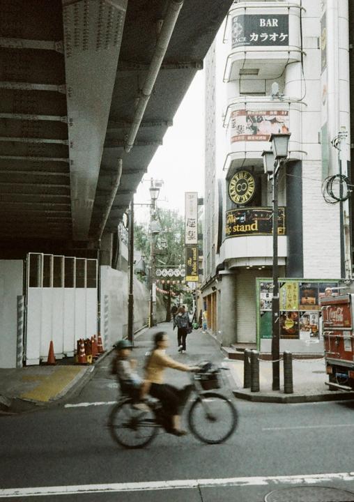 A woman cycles with a child on a city street under an overpass, with buildings and signs visible in the background.