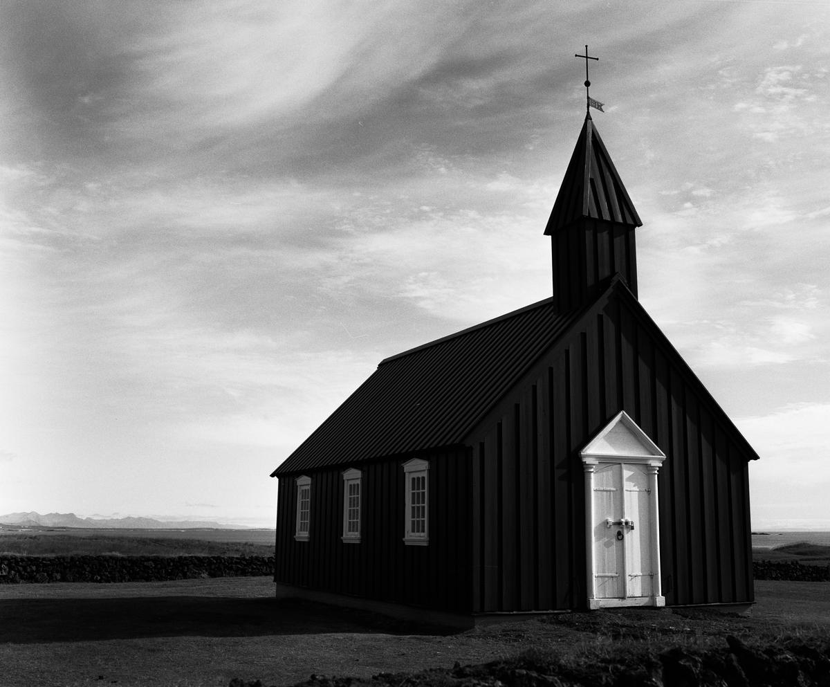 A black church with a white door and steeple stands in a field under a cloudy sky.