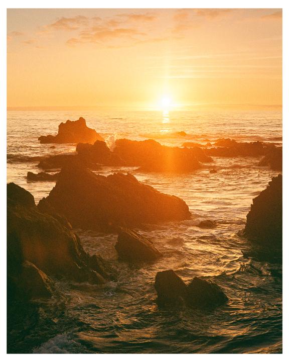 The sun sets over a rocky ocean with waves crashing against the shore.