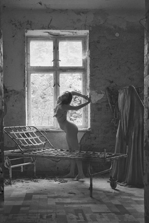 A woman stands nude in an abandoned room with a bed in the foreground, looking out a window.