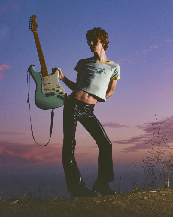 A man in a crop top and shiny pants holds a guitar against a colorful sky.