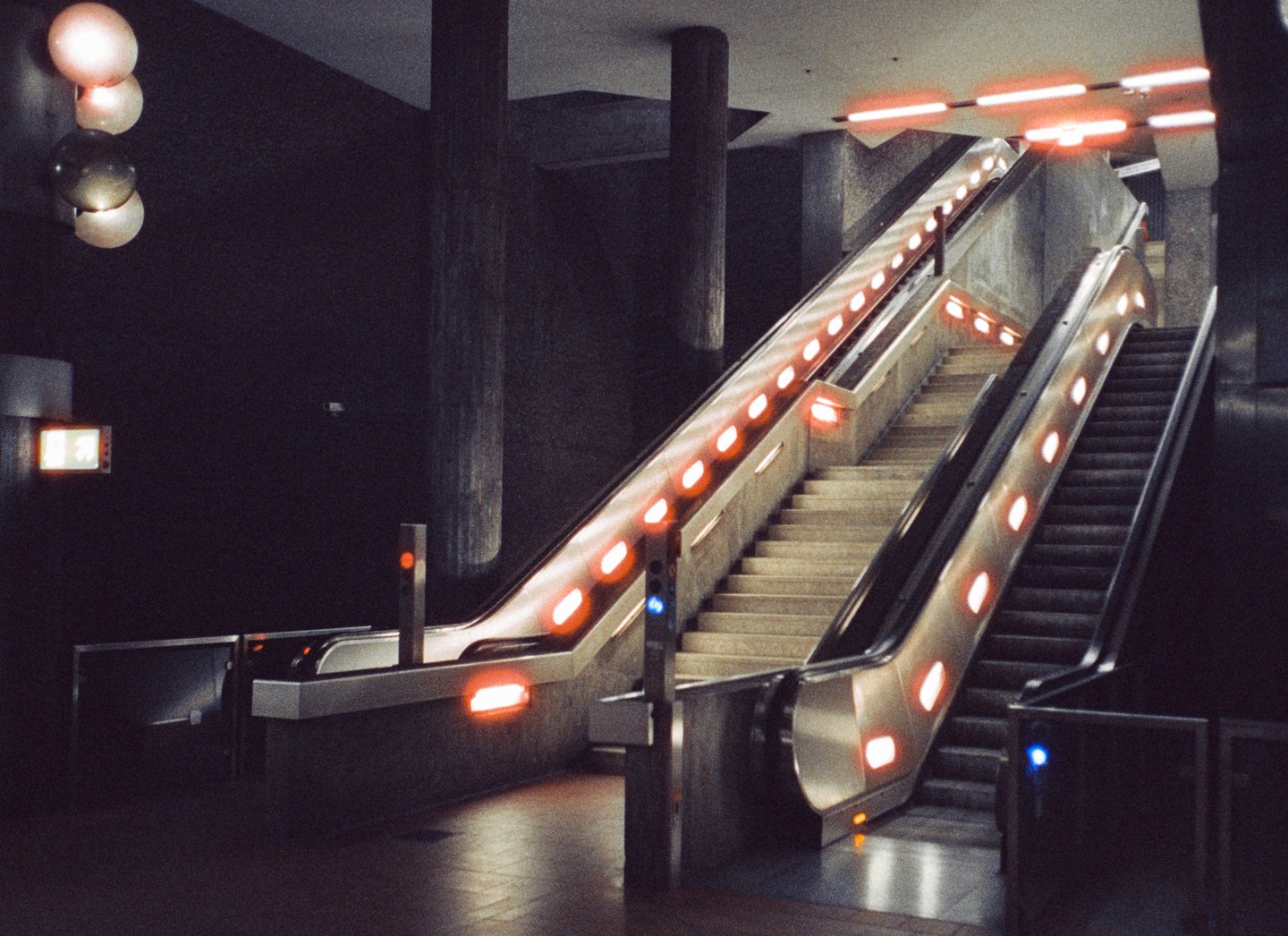 Two escalators with bright orange lights on their sides ascend from a dark subway station.
