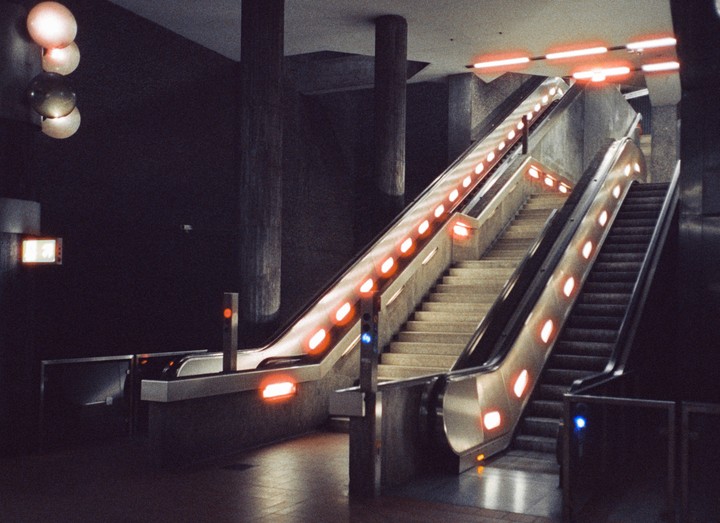 Two escalators with bright orange lights on their sides ascend from a dark subway station.
