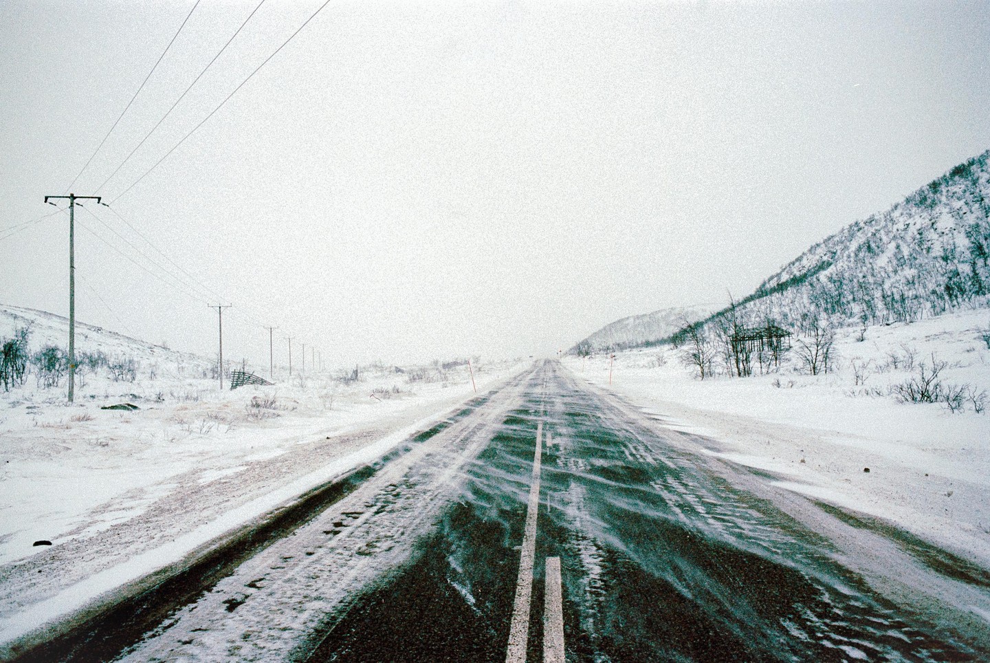A long, snow-covered road stretches into the distance, flanked by snow-covered hills and utility poles.