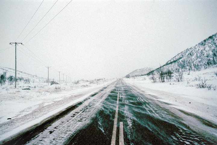 A long, snow-covered road stretches into the distance, flanked by snow-covered hills and utility poles.