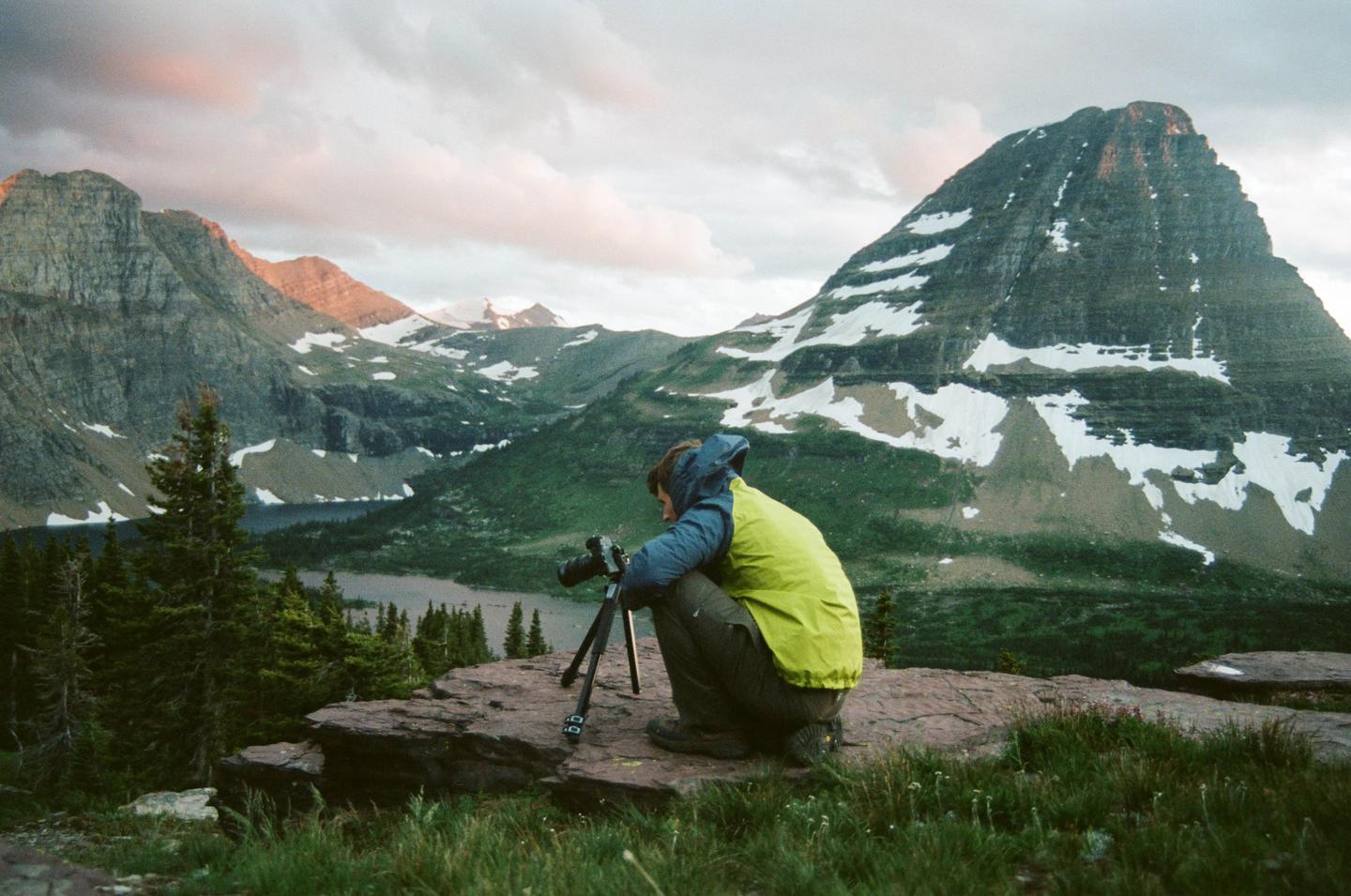 A man crouches with a camera on a tripod overlooking a lake and mountains with snow.