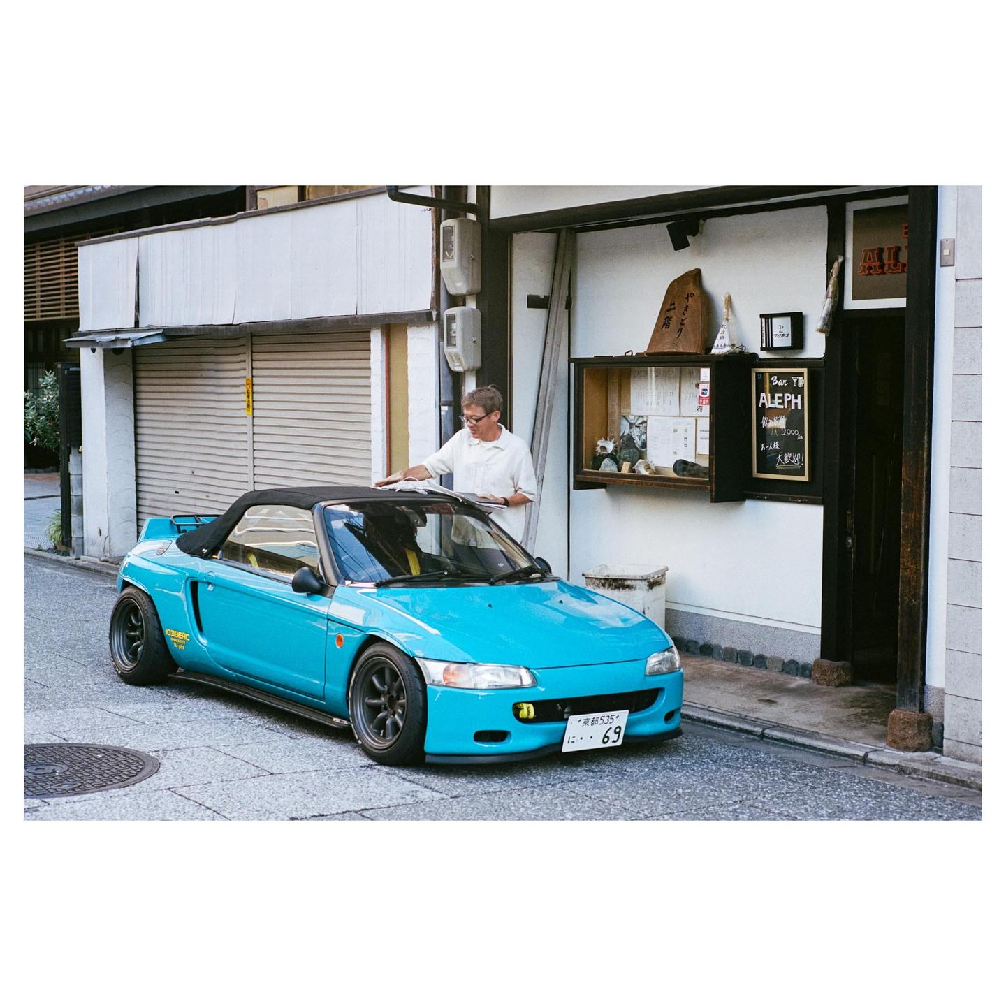 A man in a white shirt stands next to a turquoise convertible car parked on a narrow street.