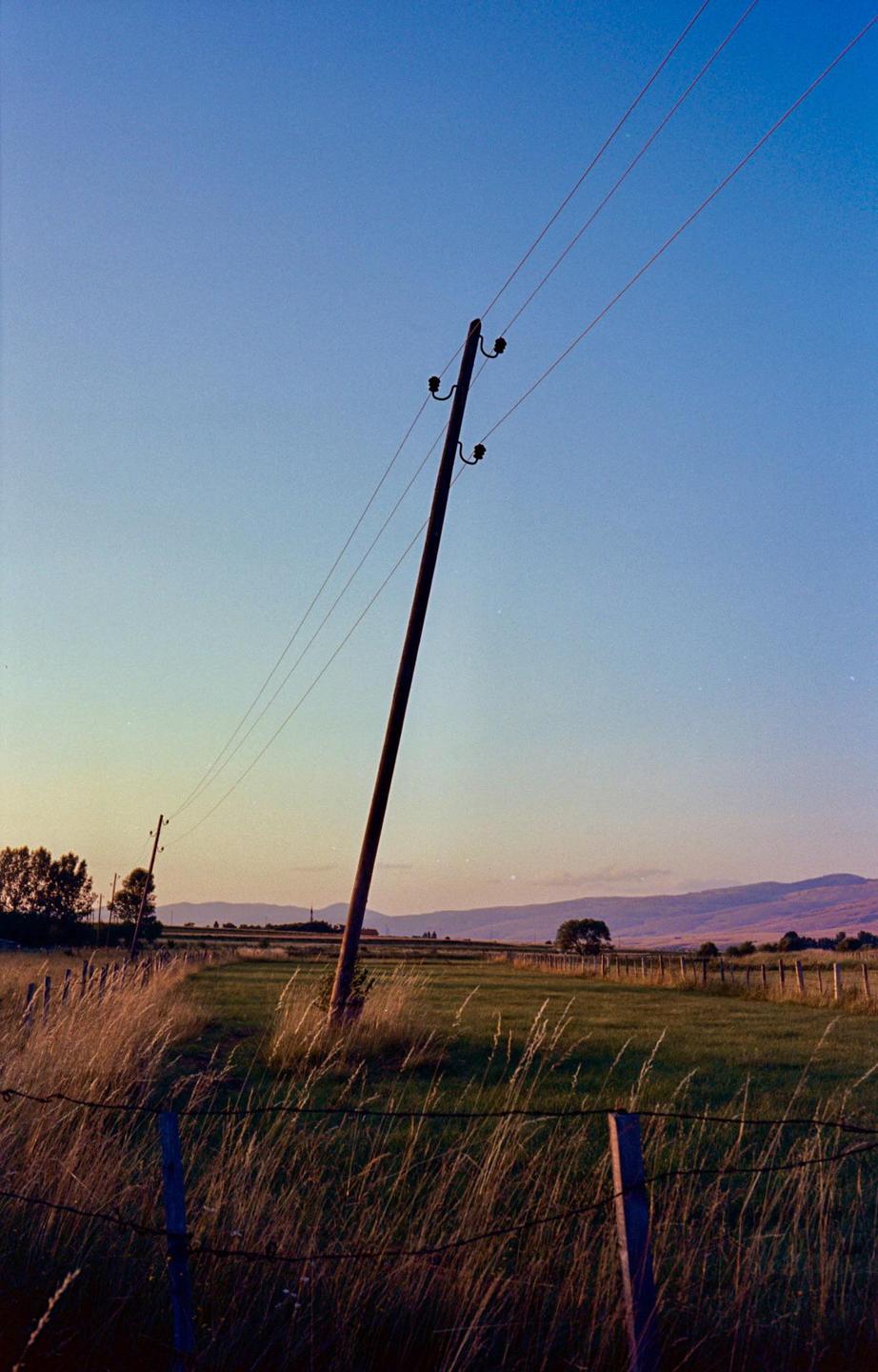 A telephone pole with wires stretches across a rural landscape with fields and mountains in the distance.