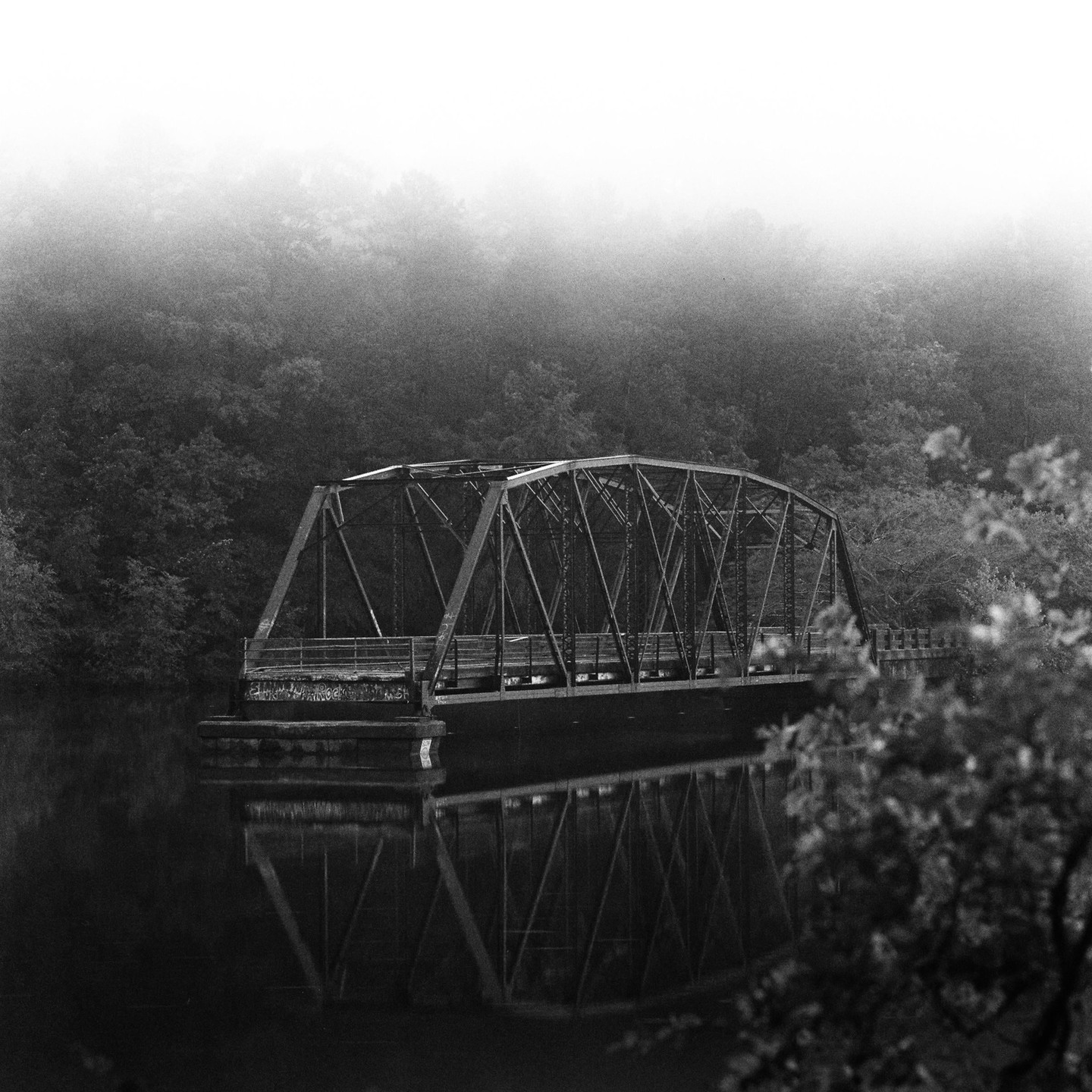 A metal truss bridge spans a body of water, reflecting on its surface, with a hazy forest in the background.