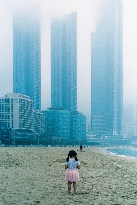 A child stands on a sandy beach looking towards the ocean and city buildings shrouded in fog.