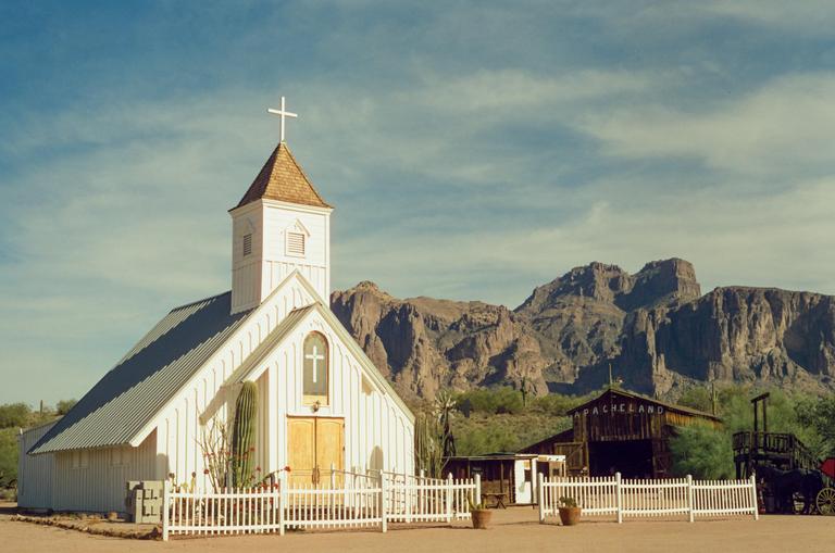 A white church with a steeple and cross stands in a desert landscape with mountains in the background.