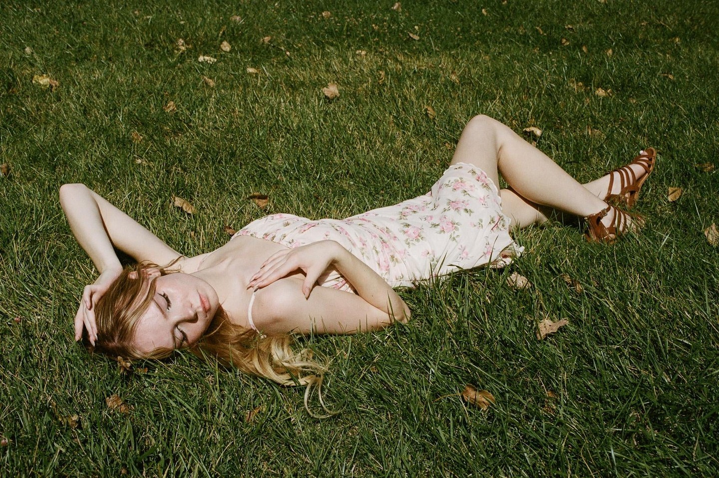 A woman in a floral dress and sandals is lying on her back in green grass with her eyes closed.