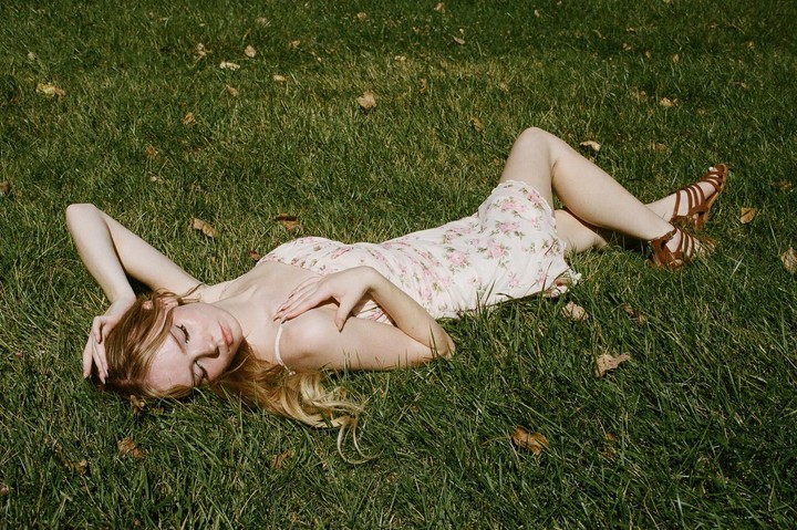 A woman in a floral dress and sandals is lying on her back in green grass with her eyes closed.