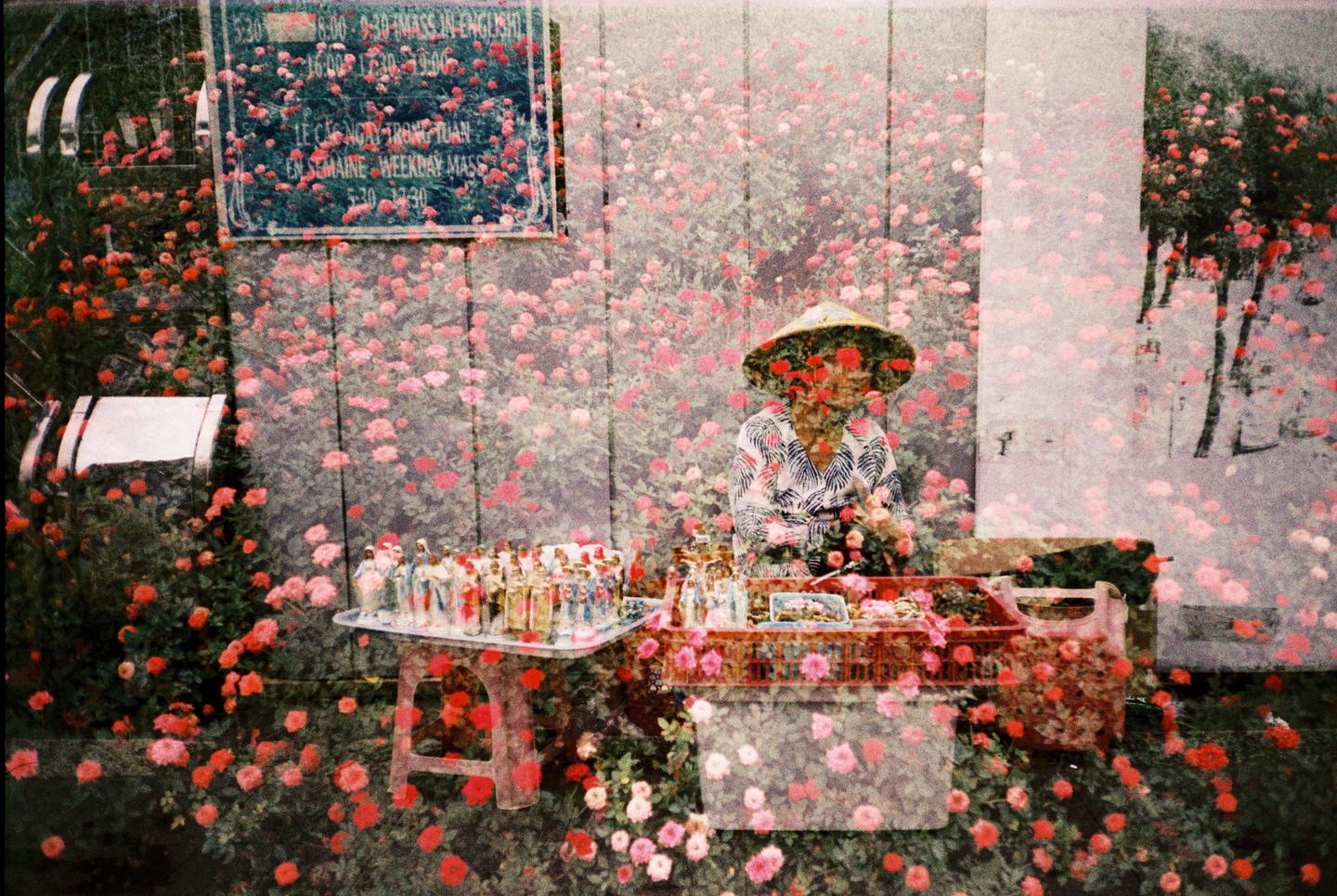 A person wearing a conical hat sits behind a stall with religious figurines and other items, all overlaid with an image of many small flowers.