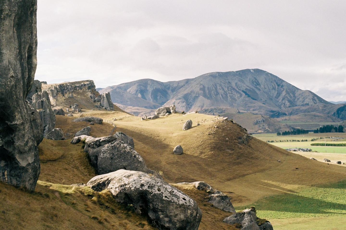 A landscape view shows large rock formations on a grassy hill with mountains in the distance under a cloudy sky.