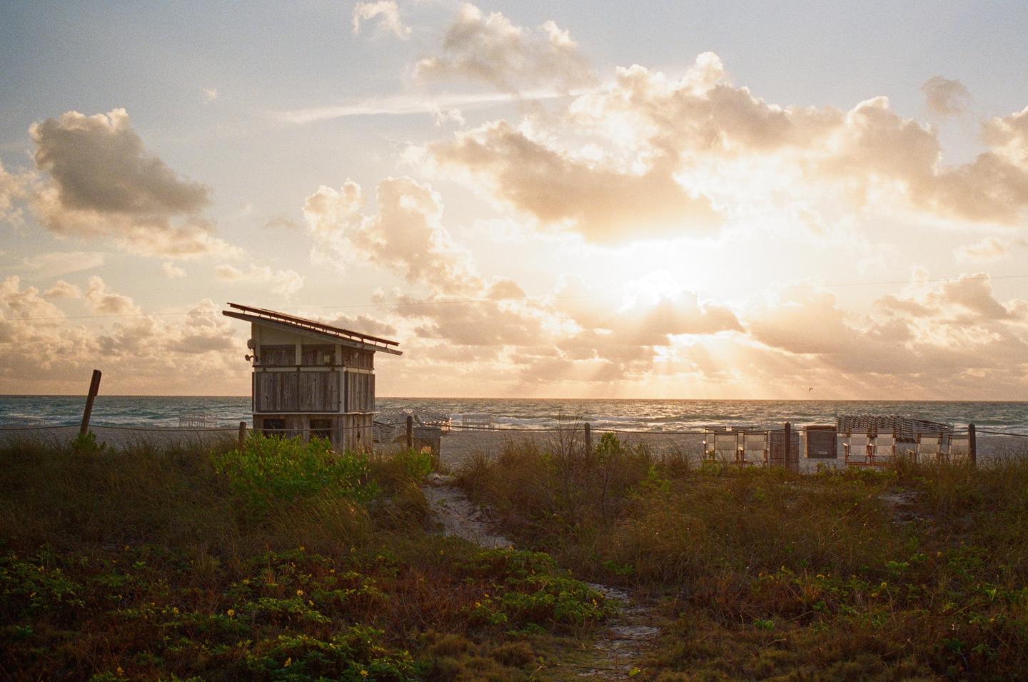 A lifeguard stand stands on a beach with the ocean in the background under a cloudy sky with sun rays.