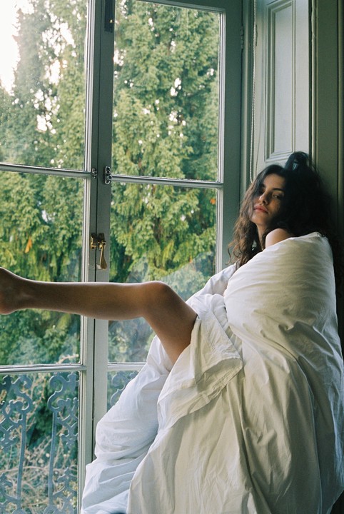 A woman wrapped in a white sheet sits by a window with her leg extended, looking towards the viewer.