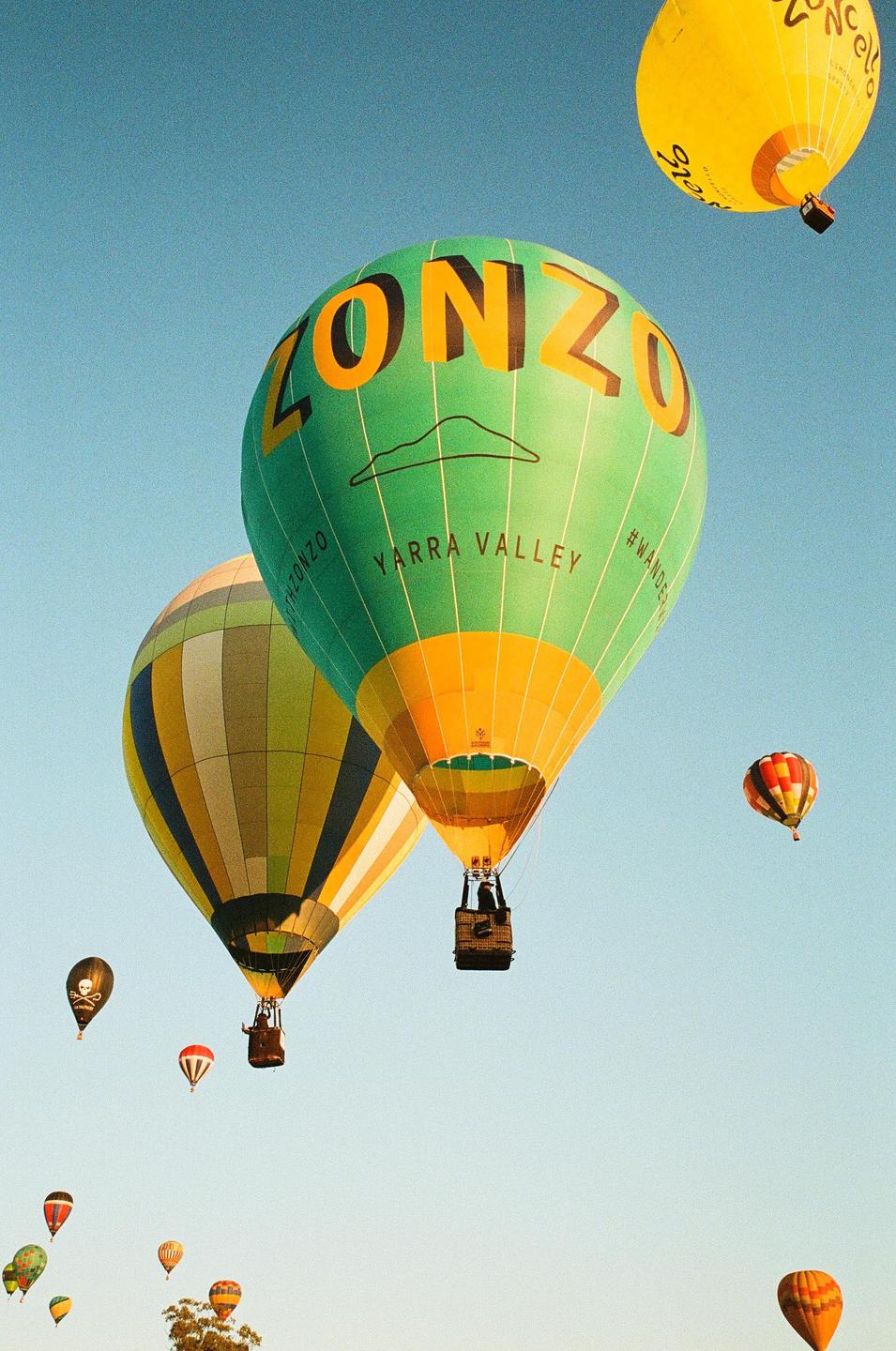 Many hot air balloons of various colors and patterns float in a clear blue sky.