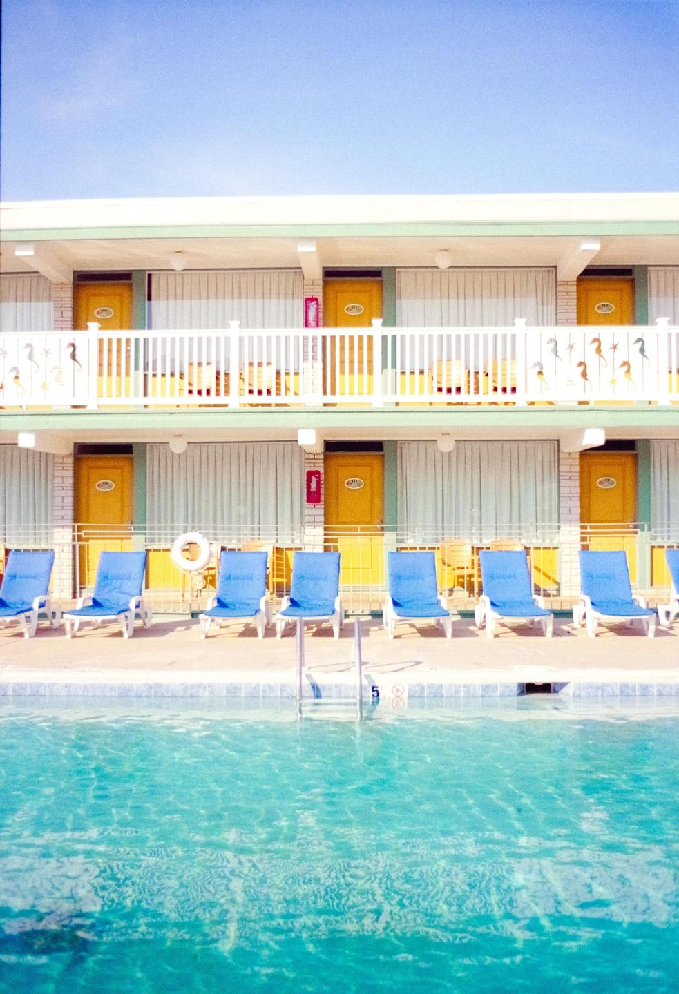 A swimming pool with blue lounge chairs in front of a two-story motel with yellow doors.