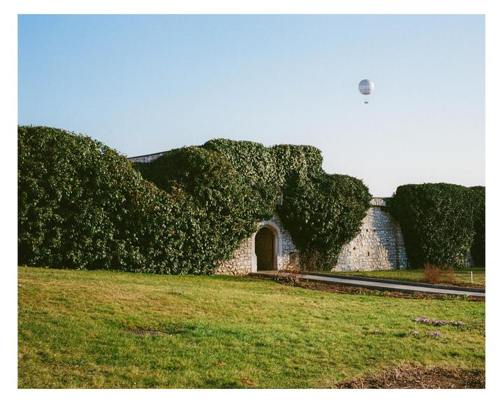 A hot air balloon floats above a stone wall covered in ivy next to a grassy field.