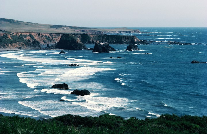 Waves crash on a rocky coastline with cliffs and hills in the background.