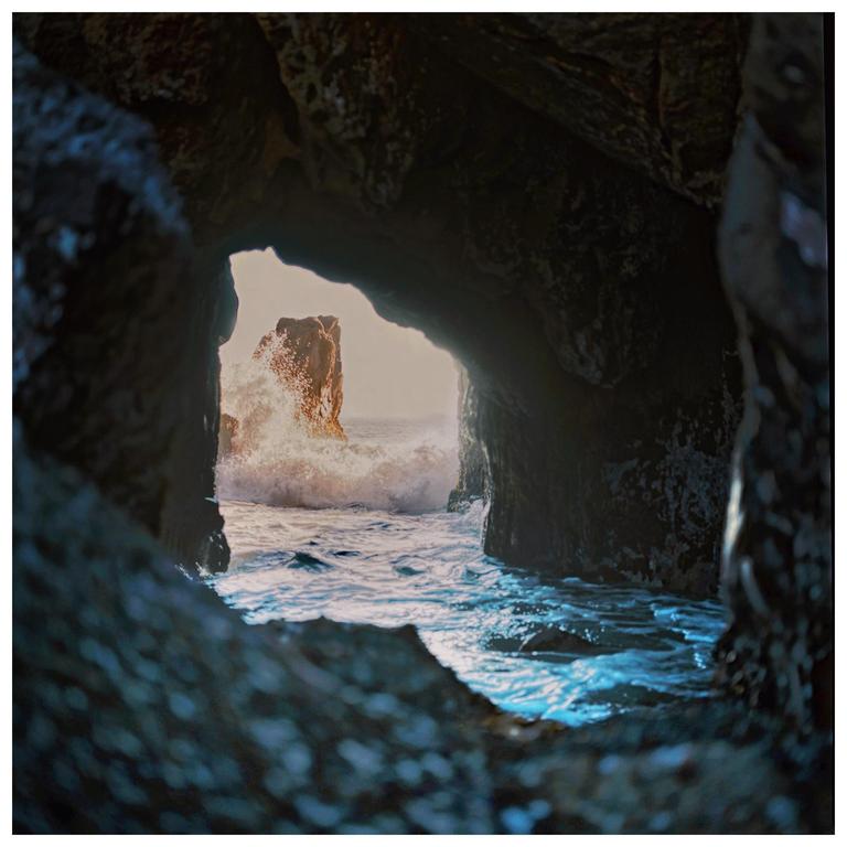 A large wave crashes against a rock formation seen through a dark cave opening.