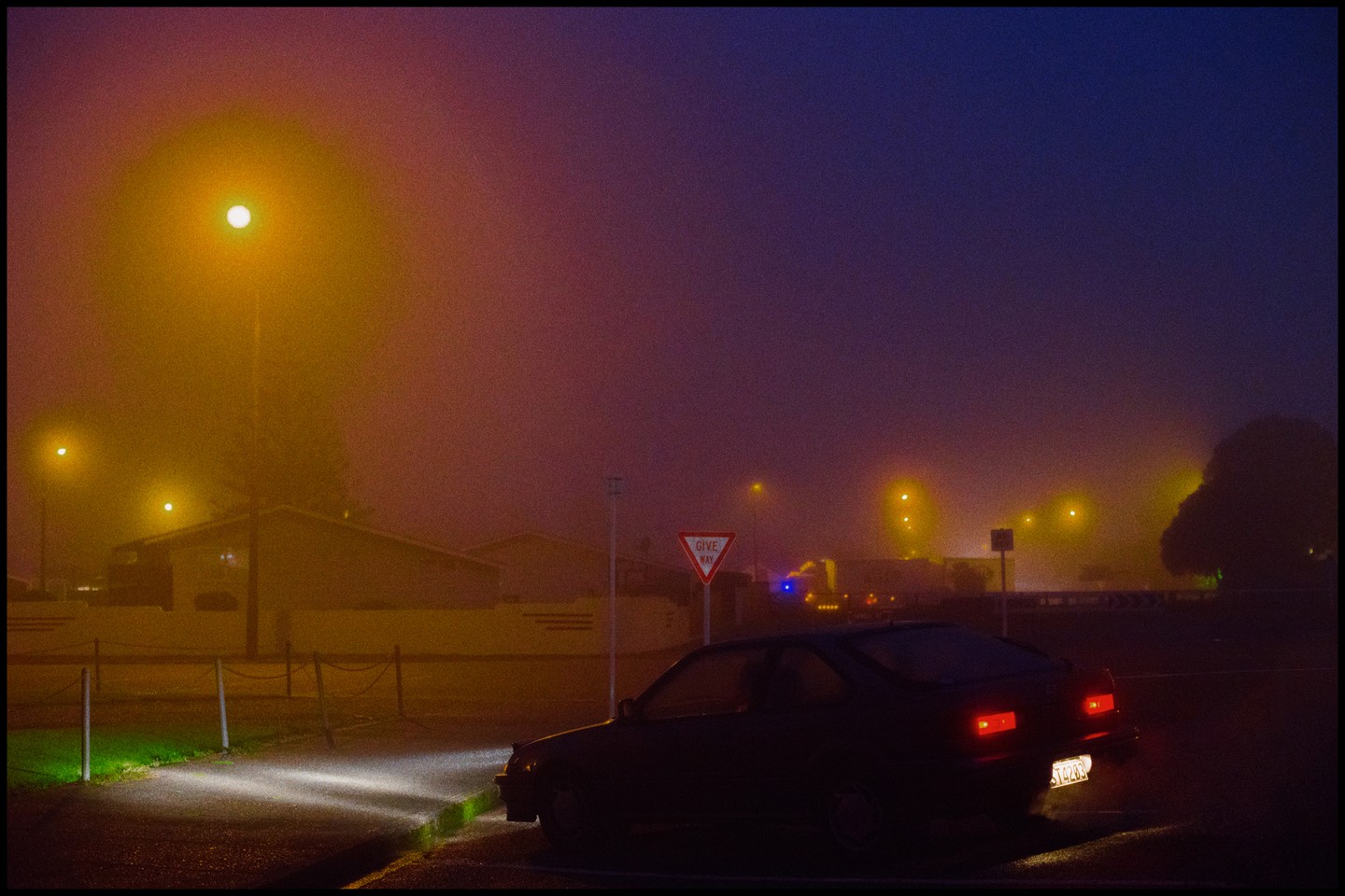 A dark car is parked on a street at night, illuminated by streetlights and its own headlights and taillights, with a "give way" sign visible in the foggy atmosphere.