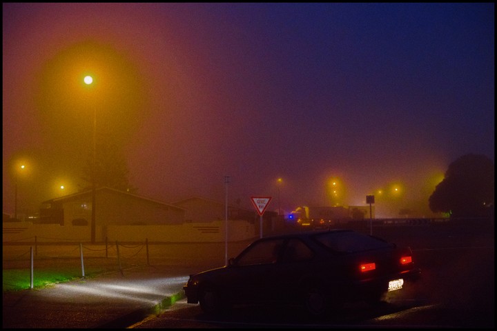 A dark car is parked on a street at night, illuminated by streetlights and its own headlights and taillights, with a "give way" sign visible in the foggy atmosphere.