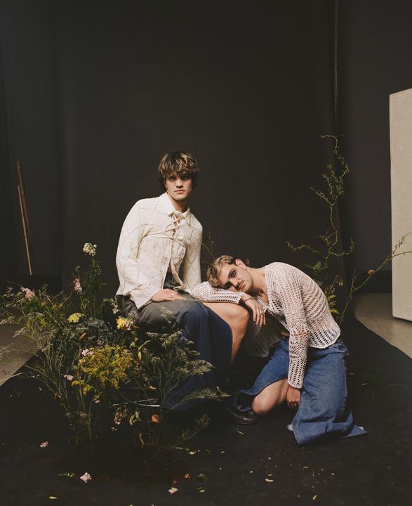 Two men pose among plants on a dark floor with a dark background.