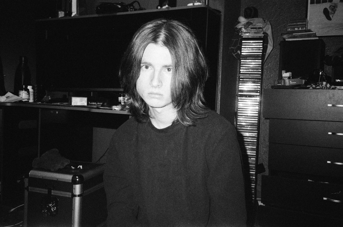 A young man with long hair looks directly at the viewer in a dimly lit room.