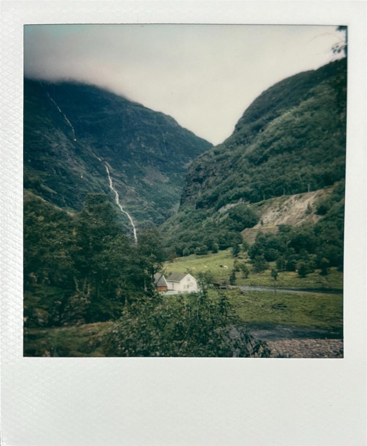 A small white house sits in a green valley with a waterfall cascading down a mountain in the background.