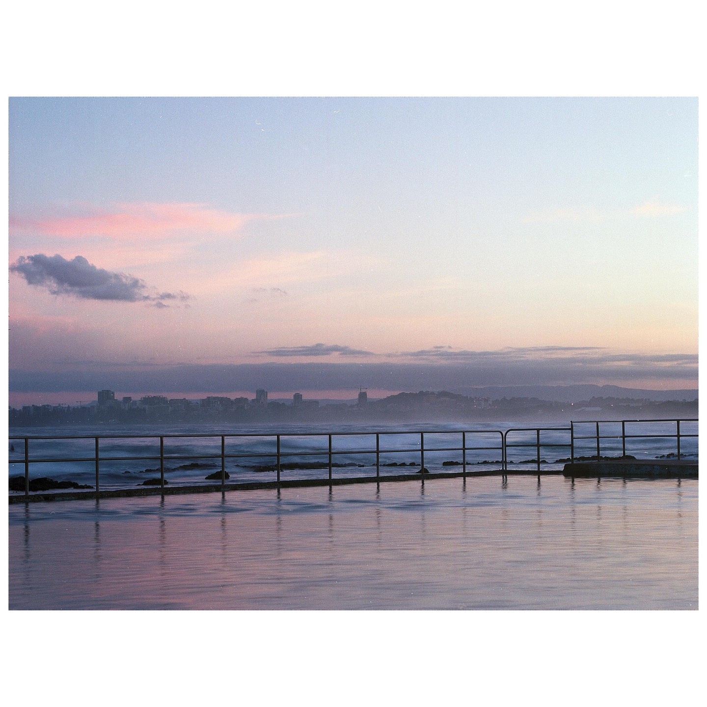 A wet concrete surface with a metal railing overlooks a city skyline and ocean at dusk.