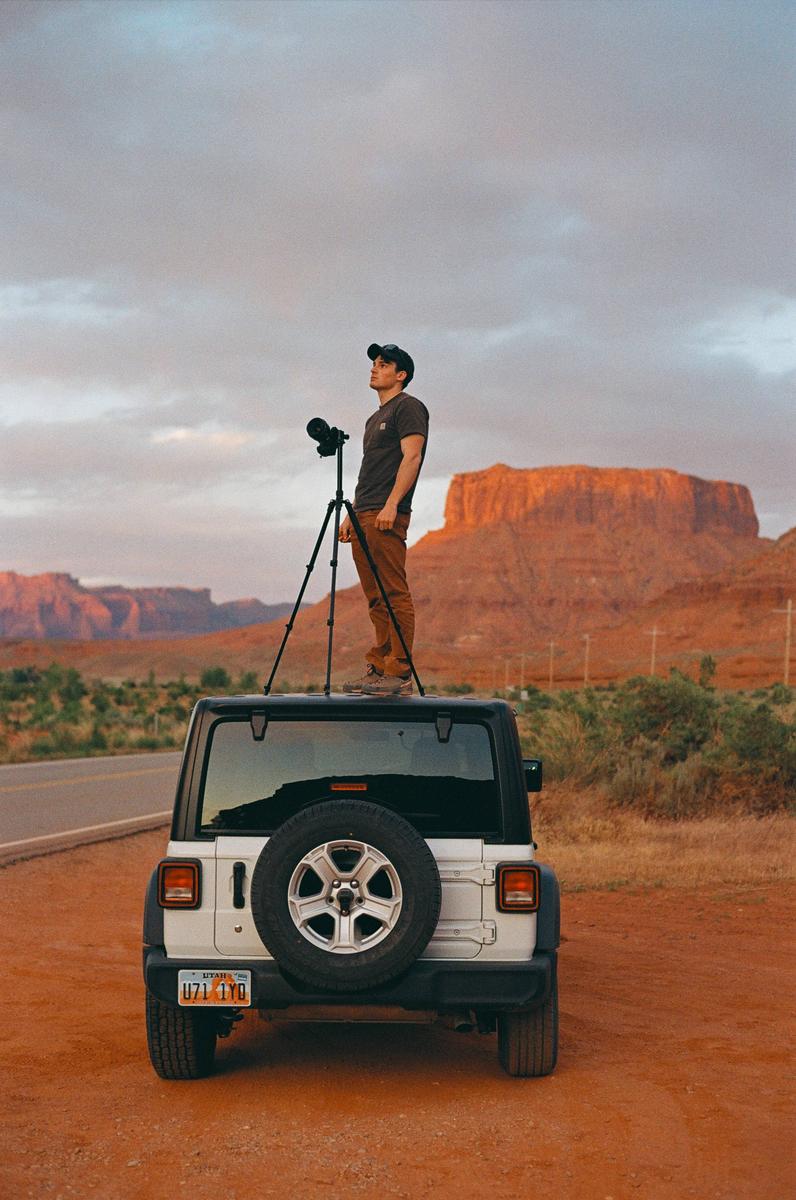 A man stands on top of a white jeep with a camera on a tripod, looking out at a desert landscape with red rock formations.