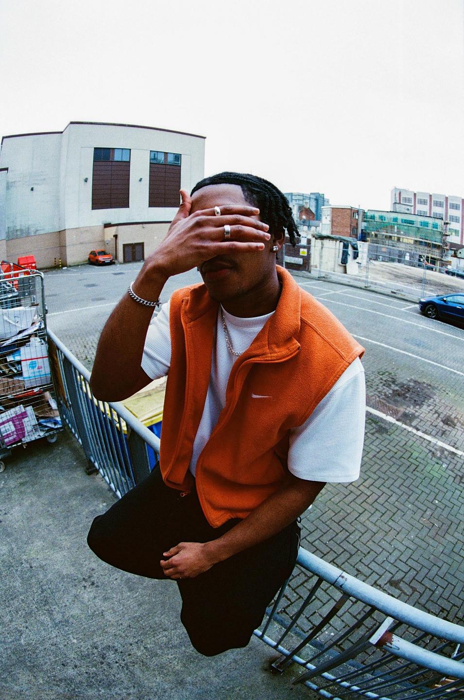 A man wearing an orange vest covers his face with his hand while sitting on a railing in an urban setting.